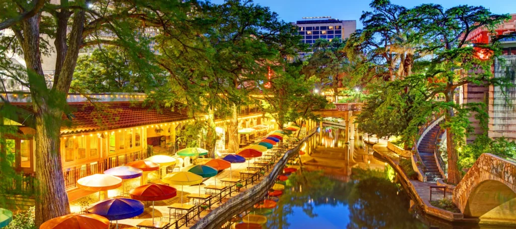 A vibrant riverside scene with colorful umbrellas over outdoor tables, lush green trees, and lit-up buildings reflecting in the calm water, likely at dusk on the San Antonio River Walk.