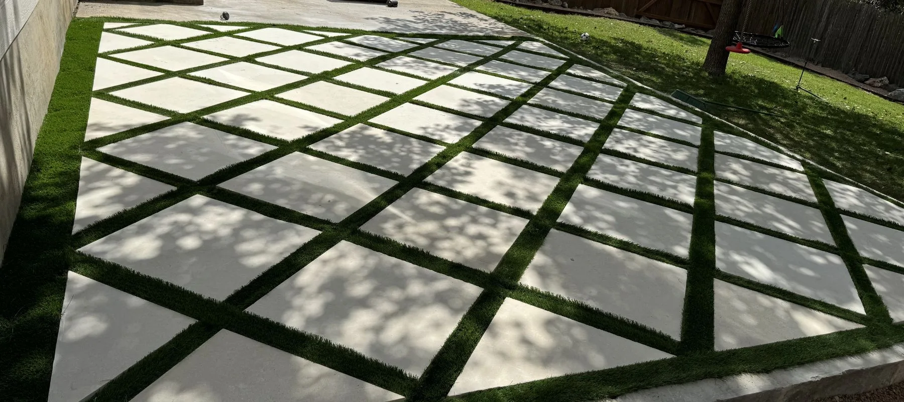 A patio features large square concrete slabs arranged in a diamond grid pattern with strips of green grass or turf between each slab, partially shaded by tree branches.