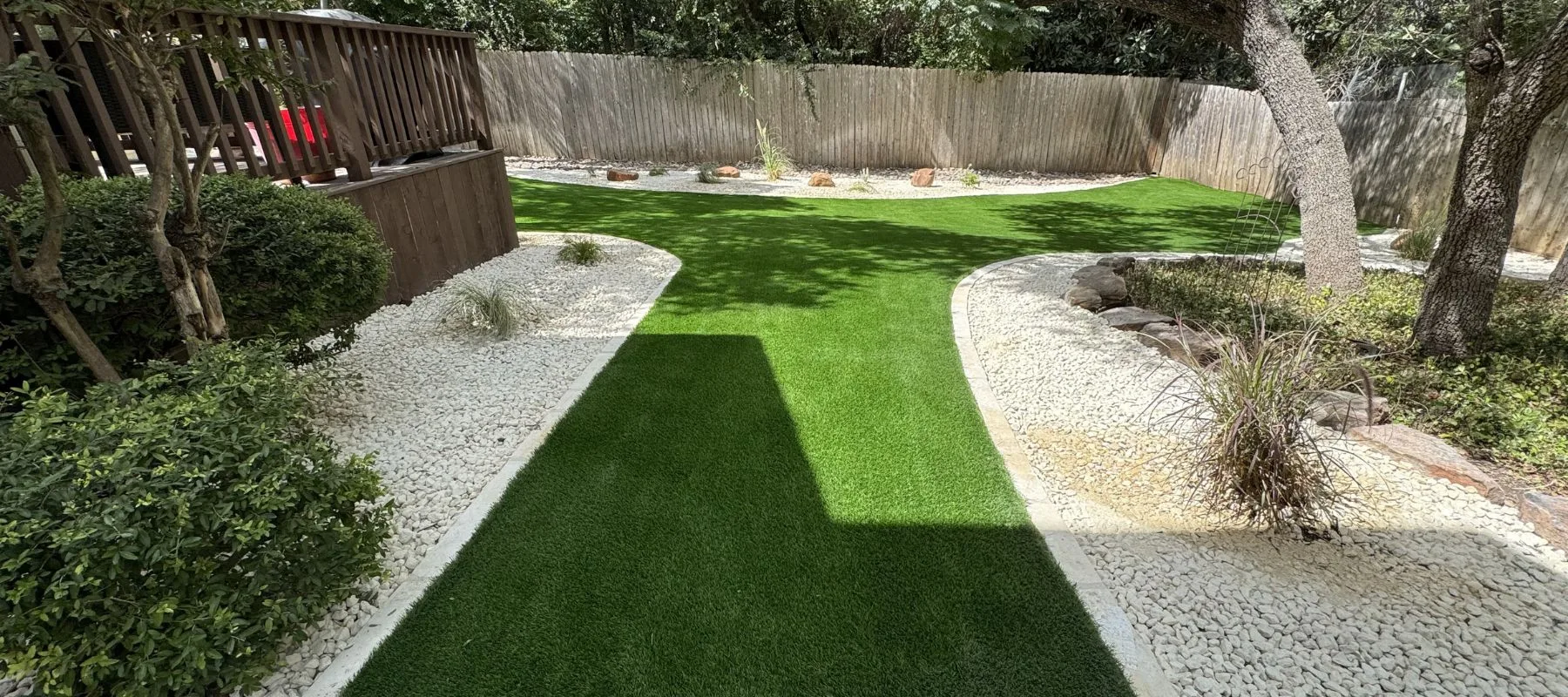 A backyard with artificial green grass, bordered by white gravel and some small plants. There are wooden fences and trees in the background, with a deck on the left side. The scene is sunny with areas of shadow.