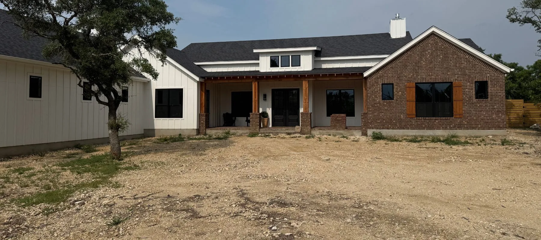 A one-story modern farmhouse with white siding and brown brick exterior, wooden shutters, a covered front porch with wooden beams, black-framed windows, and a dirt front yard with some sparse grass and trees.