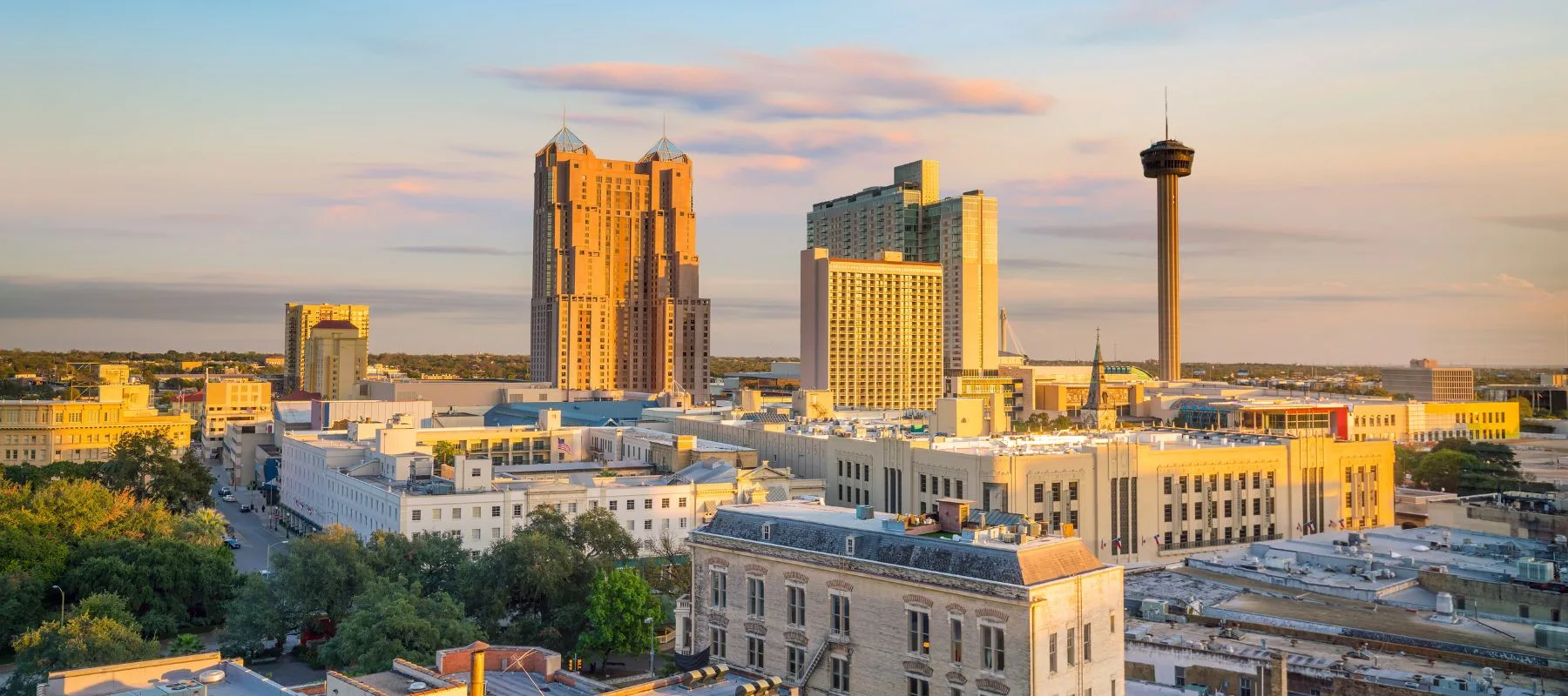A cityscape of downtown San Antonio, Texas at sunset, featuring tall buildings, the Tower Life building, and the Tower of the Americas against a blue and pink sky.