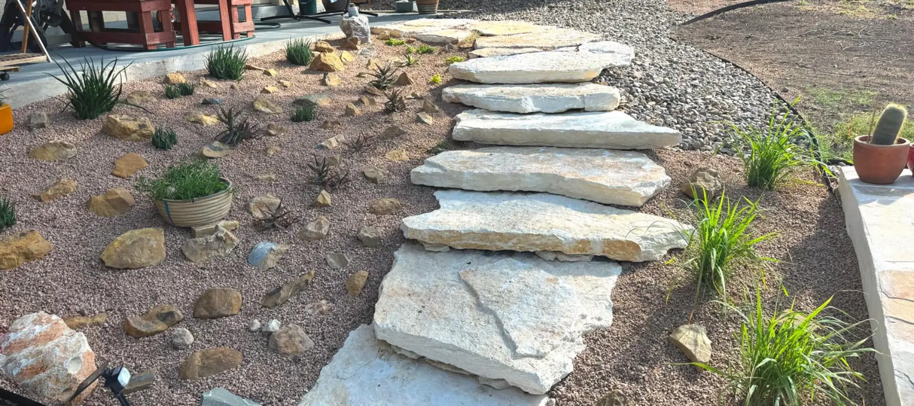 Large flat stone slabs form steps leading up a sloped garden with gravel, scattered rocks, various desert plants, and a cactus in a pot near the top right. The steps curve next to a patio on the left.