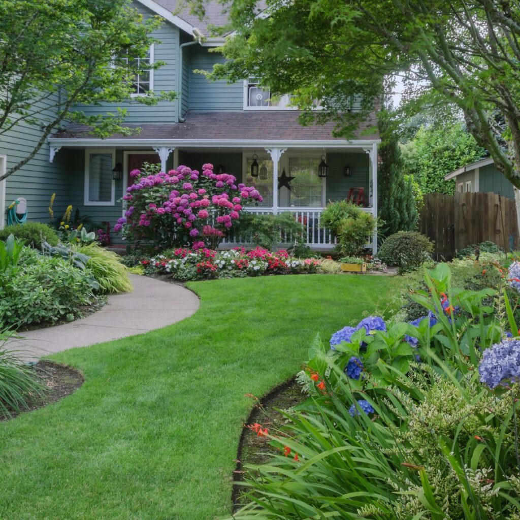 A lush, green front yard features a winding path, vibrant flower beds with pink and blue hydrangeas, and a two-story house with a covered porch and white trim surrounded by trees and shrubs.